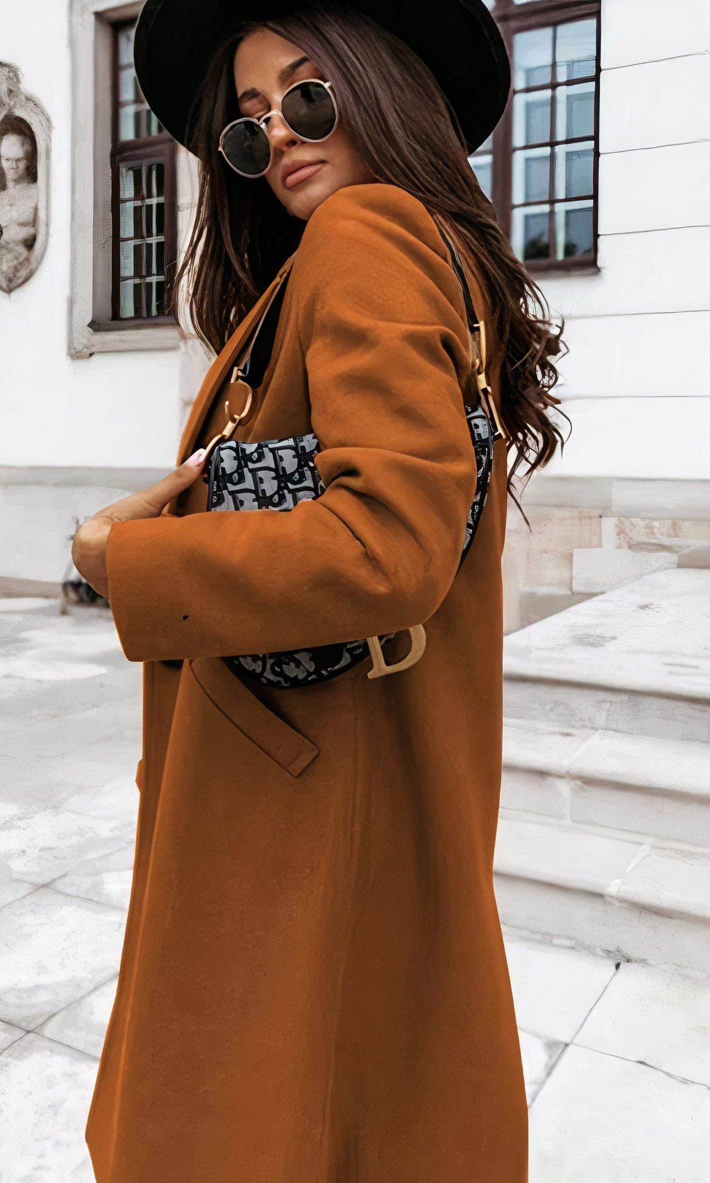 Frau in braunem Mantel und schwarzem Hut, trägt Designer-Handtasche, steht vor historischer Gebäude-Fassade. Mode, Herbst, Streetstyle.