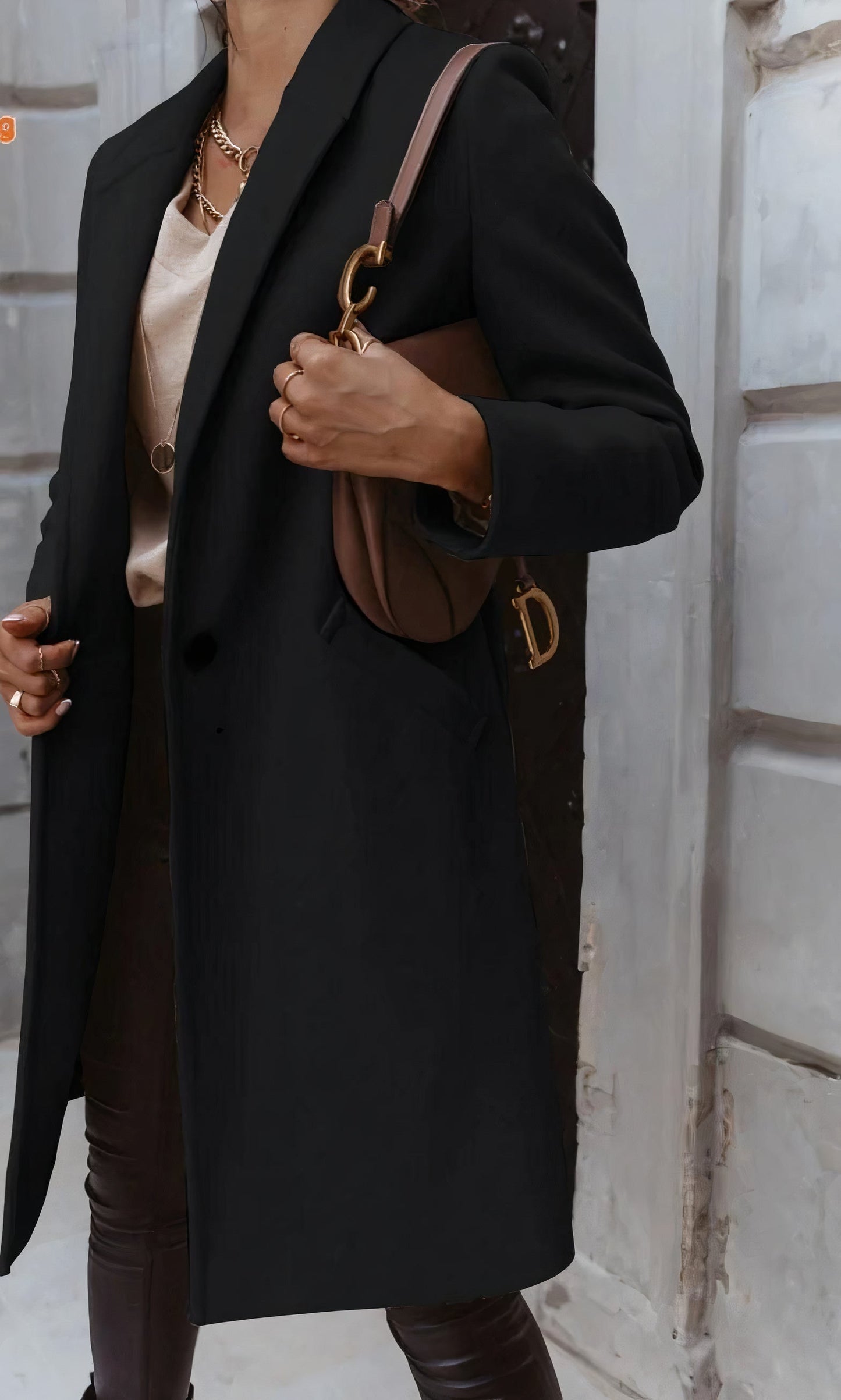 Elegante Frau in schwarzem Mantel mit brauner Designer-Handtasche, goldener Kette und beiger Bluse vor urbaner Kulisse. Fashion, Streetstyle, Mode.