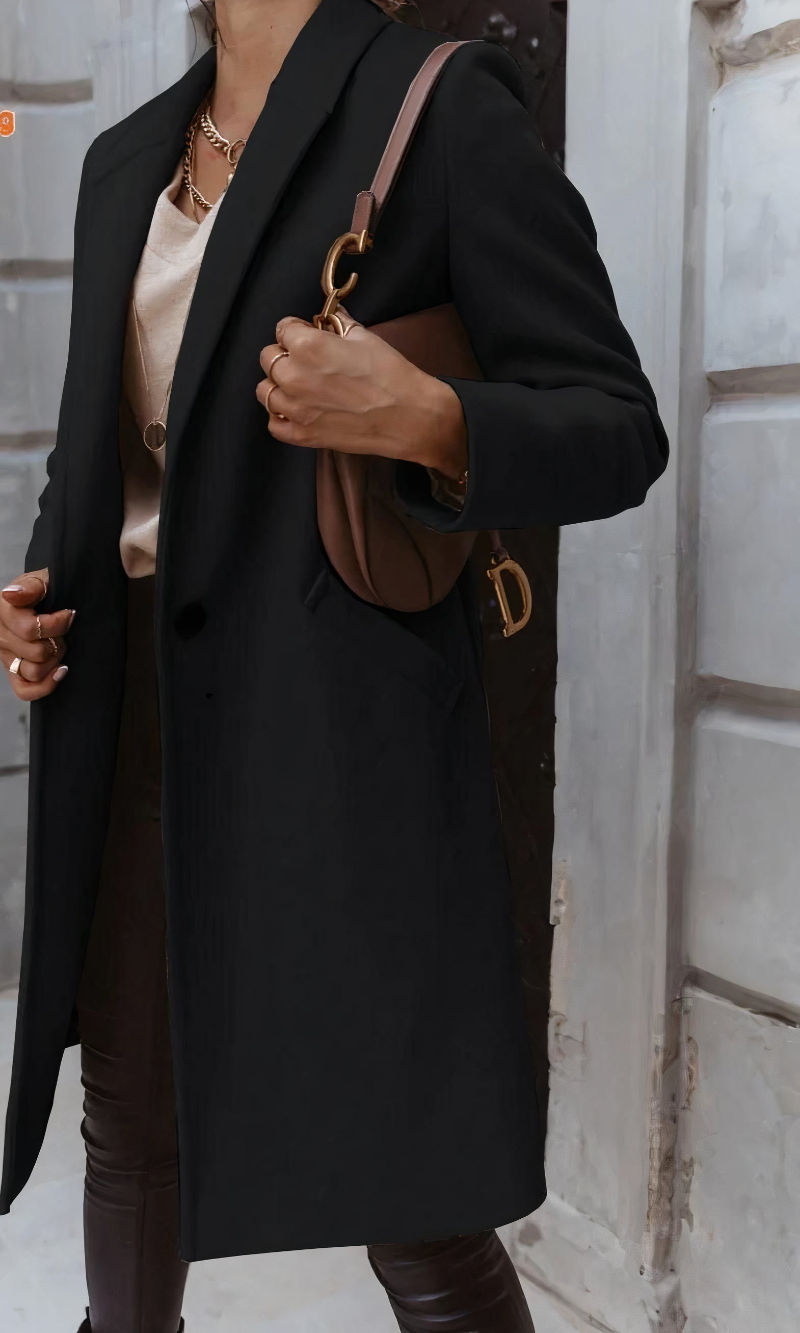Elegante Frau in schwarzem Mantel mit brauner Designer-Handtasche, goldener Kette und beiger Bluse vor urbaner Kulisse. Fashion, Streetstyle, Mode.
