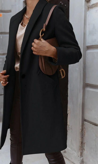 Elegante Frau in schwarzem Mantel mit brauner Designer-Handtasche, goldener Kette und beiger Bluse vor urbaner Kulisse. Fashion, Streetstyle, Mode.