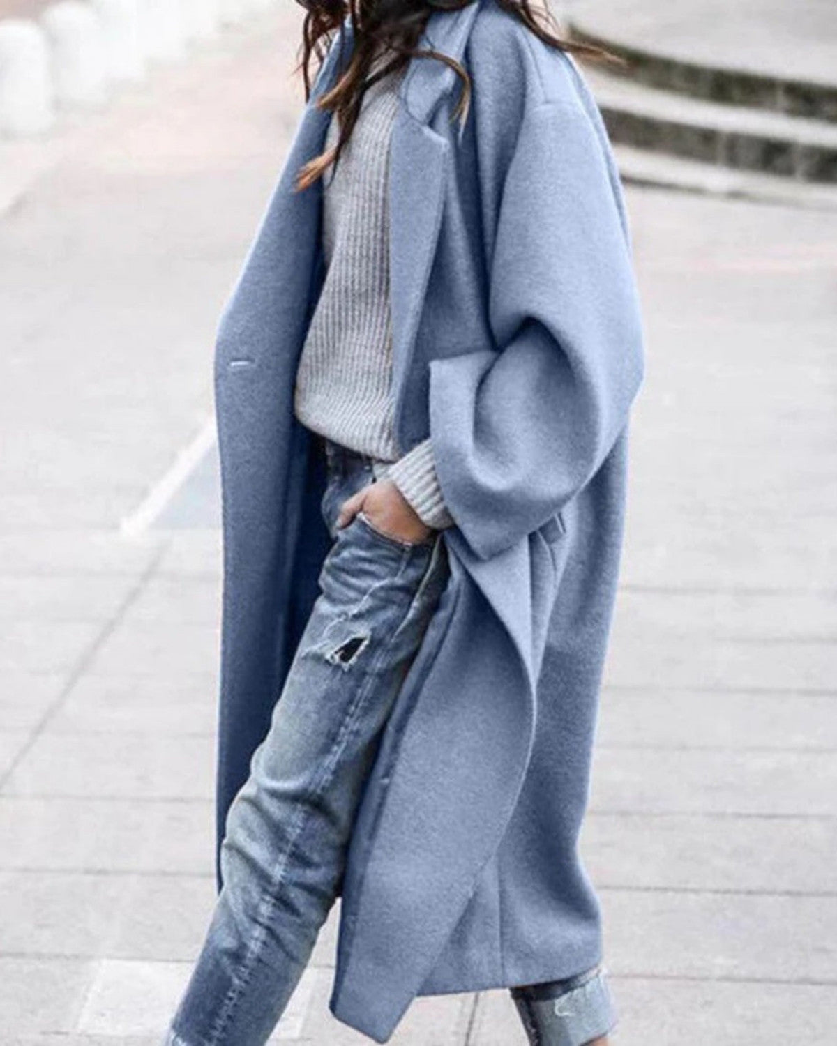 Frau in blauem Wollmantel, grauem Pullover und zerrissenen Jeans spaziert auf Gehweg. Modetrend, Herbstmode, lässiger Streetstyle.