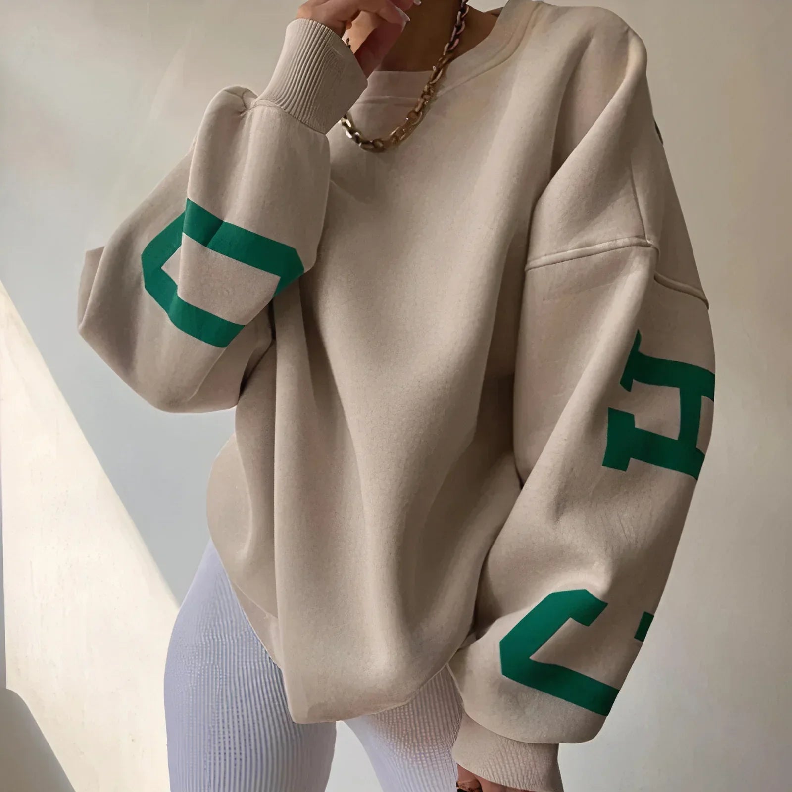 Beige Oversize-Pullover mit grünen Buchstaben auf den Ärmeln, kombiniert mit weißer Hose. Modetrend, lässiger Stil, Streetwear.