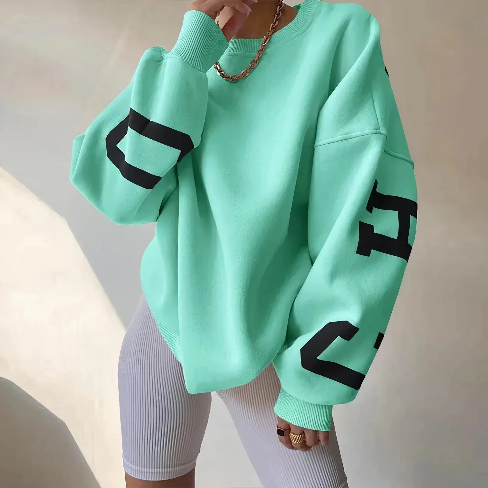 Damenmode: Oversize mintgrüner Pullover mit schwarzen Buchstaben, kombiniert mit weißen Radlerhosen. Trendiger Streetwear-Look für Herbst/Winter.