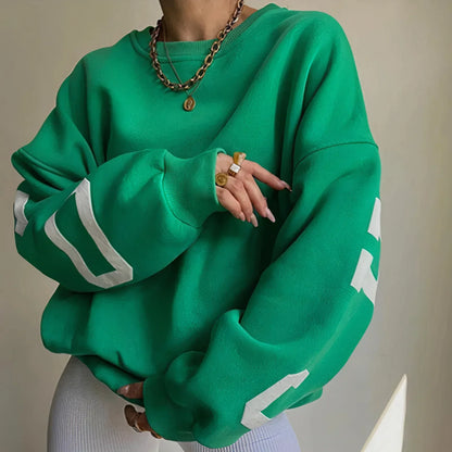 Frau in grünem Oversize-Pullover mit weißen Buchstaben, trägt goldene Ketten und Ringe. Modetrend, lässiger Stil, Streetwear.