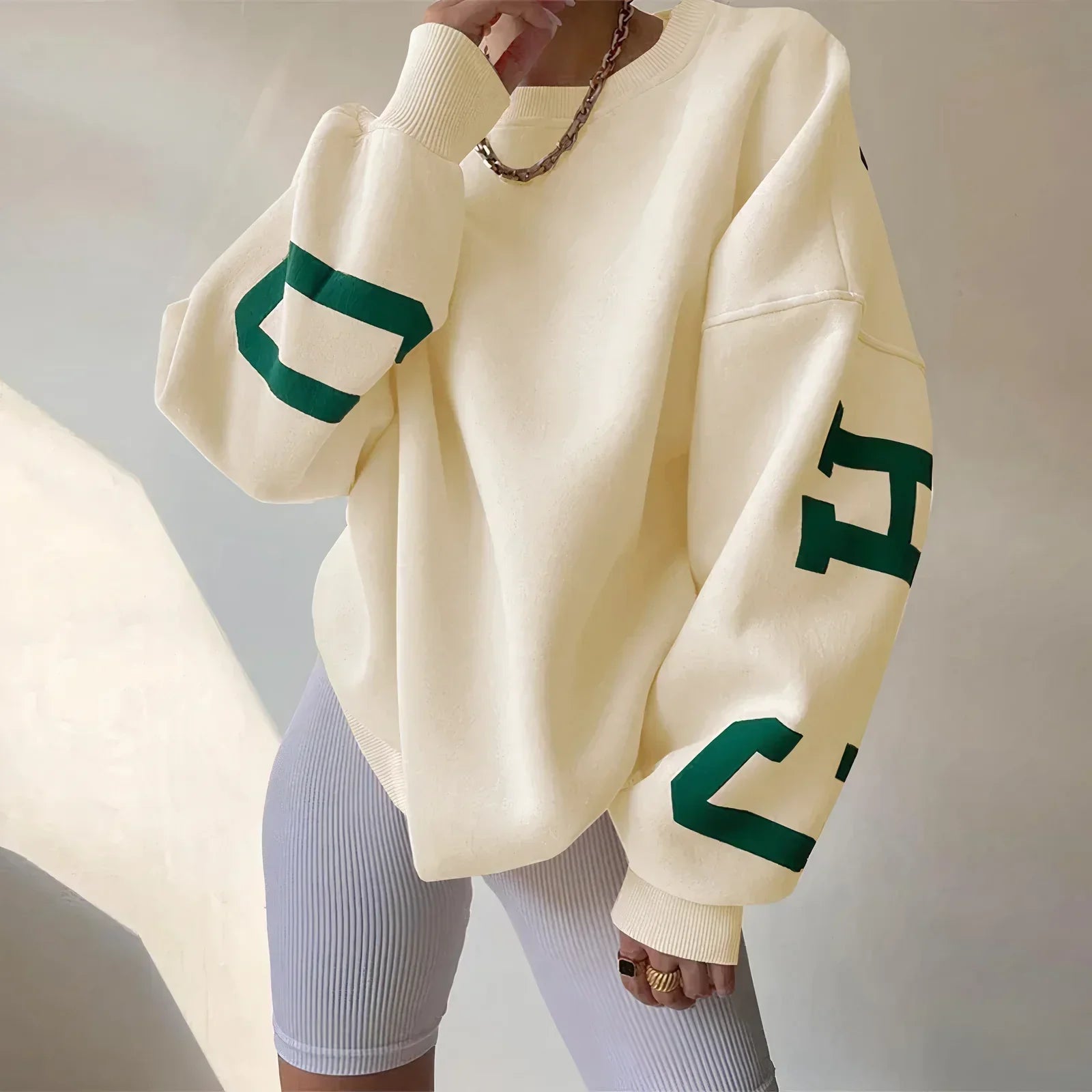 Damen Oversize Pullover in Creme mit grünen Buchstaben auf den Ärmeln, kombiniert mit weißen Radlerhosen. Modetrend, lässiger Streetstyle Look.