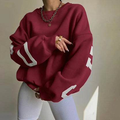 Frau in rotem Oversize-Pullover mit weißen Streifen, trägt goldene Ketten und Ringe, kombiniert mit weißen Leggings. Modetrend, lässiger Stil.