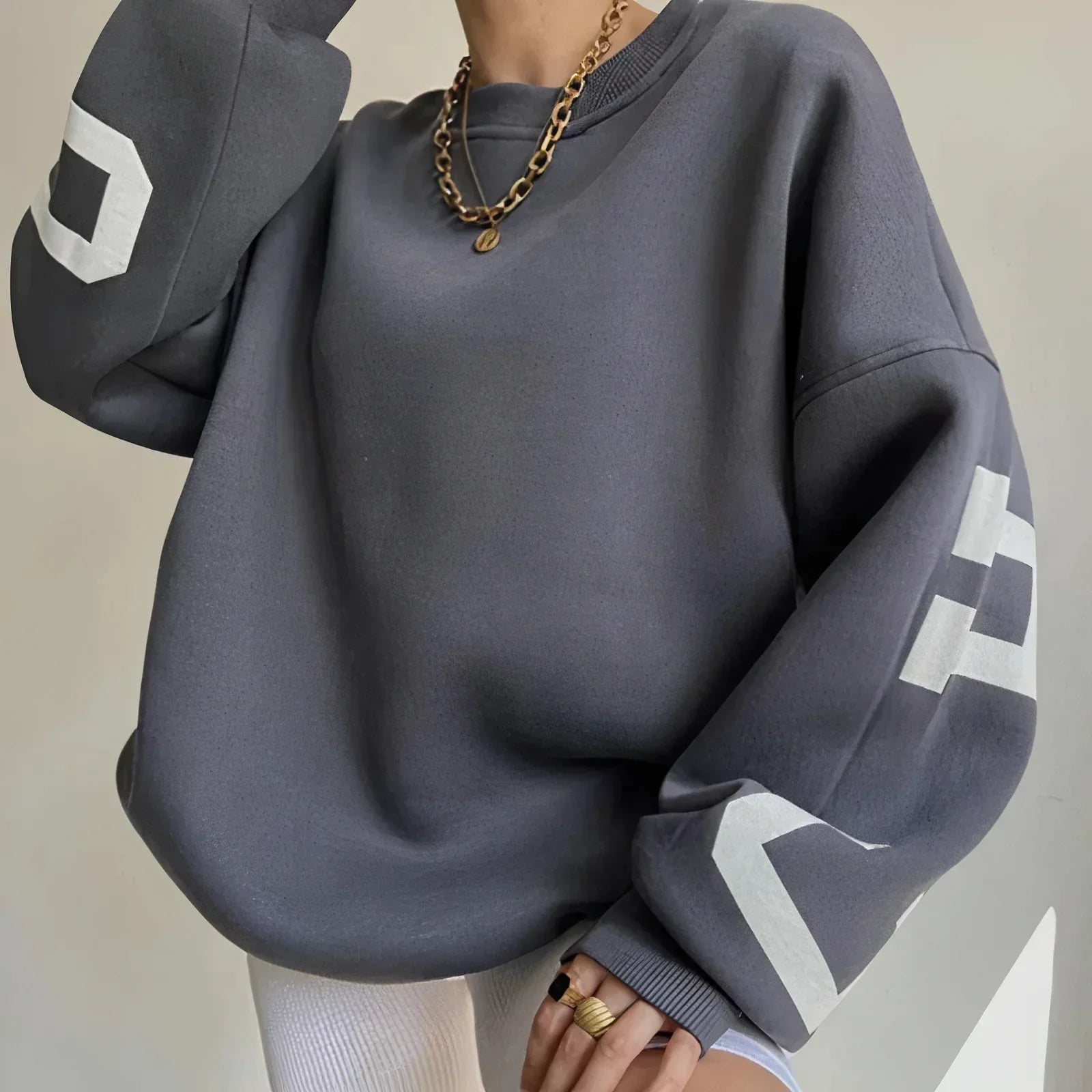 Grauer Oversize-Pullover mit weißen Buchstaben auf den Ärmeln, goldene Halskette, weiße Hose. Modetrend, lässiger Stil, Streetwear.