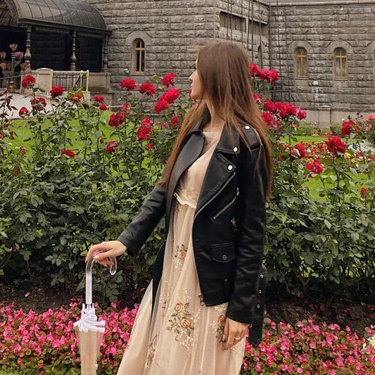 Frau in schwarzer Lederjacke und beigem Kleid steht vor einem Rosengarten, hält einen Regenschirm. Historisches Gebäude im Hintergrund.