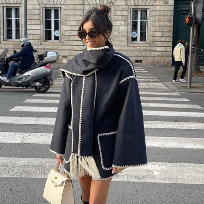 Frau in modischem, dunkelgrauem Poncho mit weißen Nähten, Sonnenbrille und Handtasche überquert Straße in städtischer Umgebung.