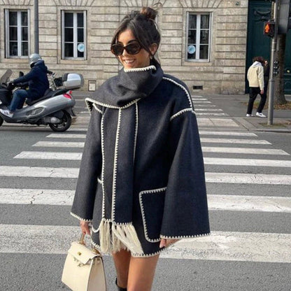 Frau in modischem, dunkelgrauem Poncho mit weißen Nähten, Sonnenbrille und Handtasche überquert Straße in städtischer Umgebung.