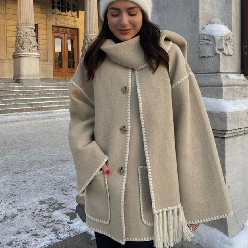 Frau in beigem Wollmantel mit Kapuze und Fransen, steht vor einem historischen Gebäude im Winter. Warme Wintermode, stilvoller Mantel, Stadtbummel.