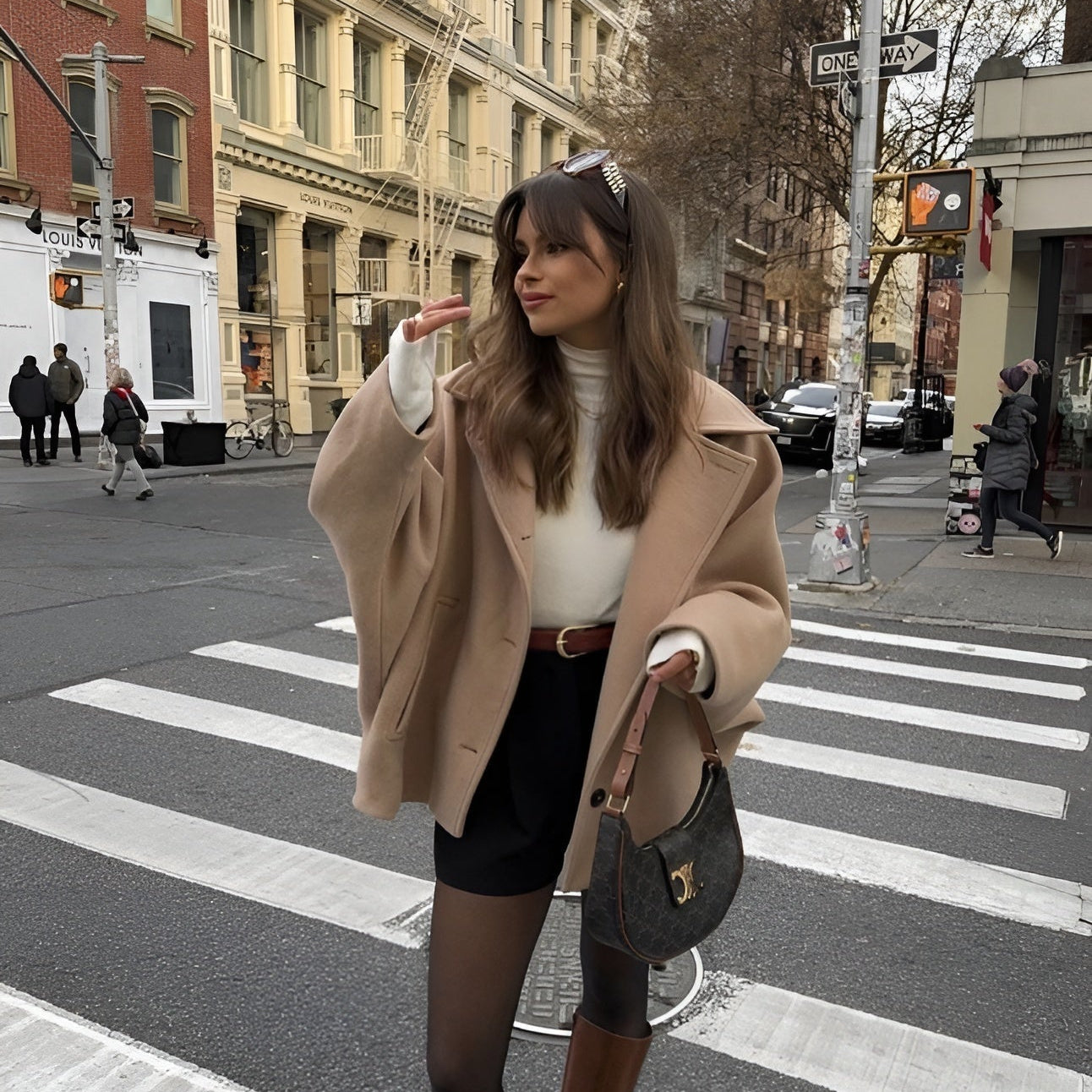 Frau im beigen Mantel und schwarzem Rock überquert eine Straße in der Stadt. Sie trägt eine Handtasche und posiert auf einem Zebrastreifen.