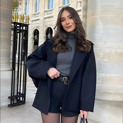 Eine Frau in eleganter schwarzer Kleidung steht vor einem historischen Gebäude. Sie trägt einen Mantel, Rollkragenpullover und Shorts.