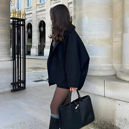 Frau in schwarzem Mantel mit großer schwarzer Handtasche vor historischer Architektur. Elegante Mode, Herbstoutfit, Stadtbummel.
