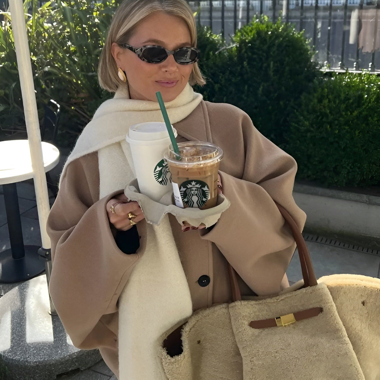 Frau mit Sonnenbrille hält zwei Starbucks-Getränke, trägt beigen Mantel und weiße Schal, sitzt im Freien. Trendige Herbstmode, Kaffee, Lifestyle.