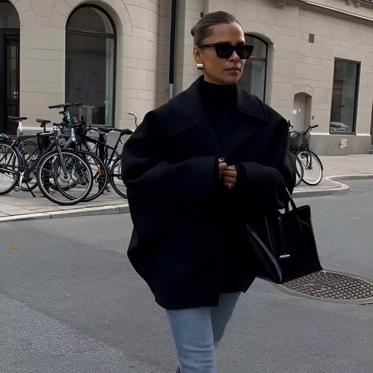 Frau in übergroßem schwarzem Mantel und Sonnenbrille, trägt schwarze Handtasche, geht auf städtischer Straße mit Fahrrädern im Hintergrund.