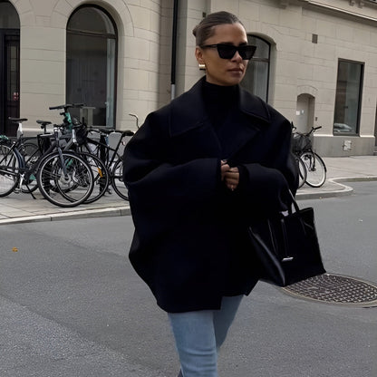 Frau in übergroßem schwarzem Mantel und Sonnenbrille, trägt schwarze Handtasche, geht auf städtischer Straße mit Fahrrädern im Hintergrund.