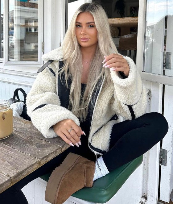 Blonde Frau in weißer Teddyjacke sitzt in Café, hält Getränk. Modetrend, Wintermode, gemütlicher Look, Outdoor-Café, lässiger Stil.