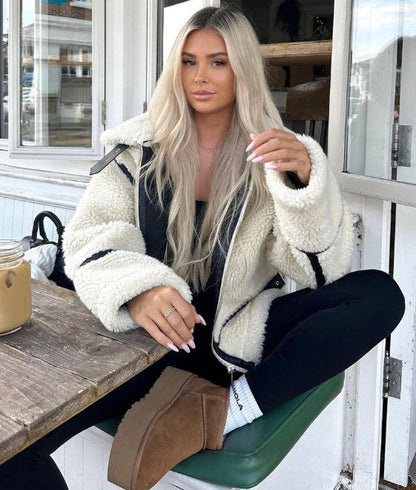 Blonde Frau in weißer Teddyjacke sitzt in Café, hält Getränk. Modetrend, Wintermode, gemütlicher Look, Outdoor-Café, lässiger Stil.