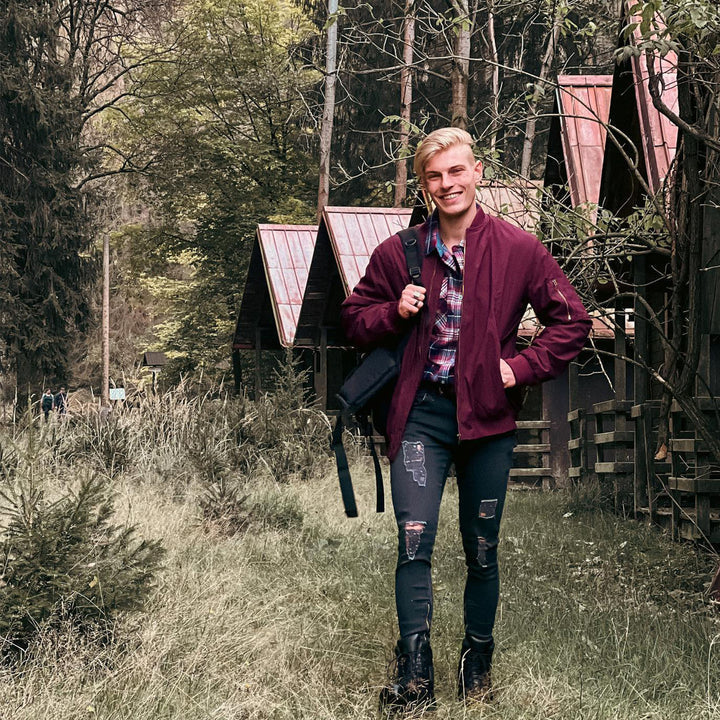Ein lächelnder Mann in roter Jacke und zerrissenen Jeans spaziert auf einem Waldweg vor Holzhütten. Natur, Outdoor, Freizeit, Waldspaziergang.