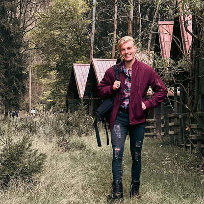 Ein lächelnder Mann in roter Jacke und zerrissenen Jeans spaziert auf einem Waldweg vor Holzhütten. Natur, Outdoor, Freizeit, Waldspaziergang.