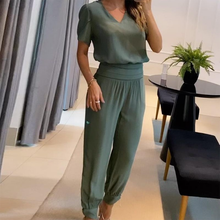 Frau in eleganter, grüner Bluse und Hose, steht in modernem Wohnzimmer mit Tisch und Pflanze. Stilvolle Damenmode, Freizeitoutfit, Inneneinrichtung.