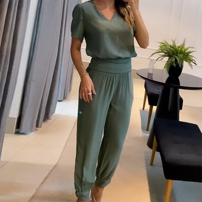 Frau in eleganter, grüner Bluse und Hose, steht in modernem Wohnzimmer mit Tisch und Pflanze. Stilvolle Damenmode, Freizeitoutfit, Inneneinrichtung.
