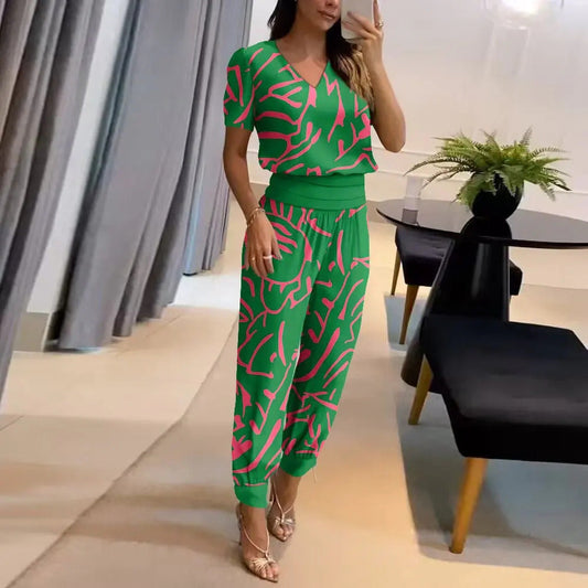 Frau in grünem, gemustertem Jumpsuit posiert in modernem Wohnzimmer mit Pflanzen und schwarzem Tisch. Elegante Mode, Innenarchitektur, Stil.