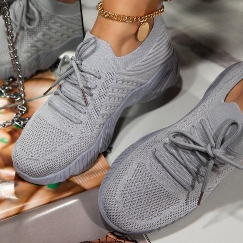 Graue Damen-Sneaker mit Strickmuster und Schnürsenkeln, getragen mit goldener Fußkette. Modische, bequeme Schuhe für Freizeit und Sport.