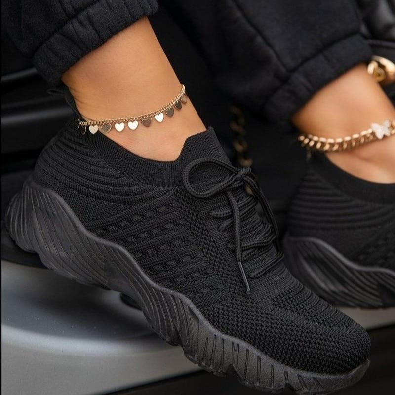 Schwarze Strick-Sneaker mit dicker Sohle, getragen von Person mit Herzfußkettchen. Modische Damen-Schuhe, bequem und trendig.
