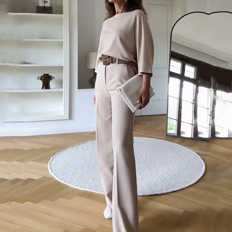 Frau in eleganter, beiger Hose und Oberteil, steht in modernem Wohnzimmer mit Parkettboden, rundem Teppich und großem Spiegel.