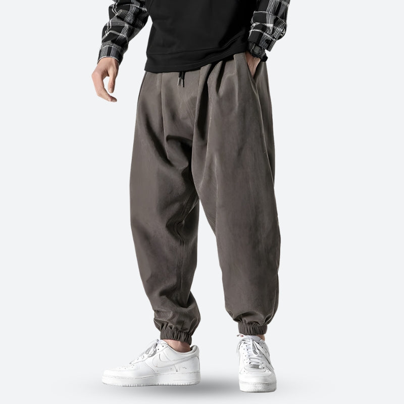Mann trägt graue, weite Haremshose mit elastischen Bündchen, kombiniert mit weißem Sneaker und kariertem Hemd. Modetrend, lässiger Streetwear-Look.