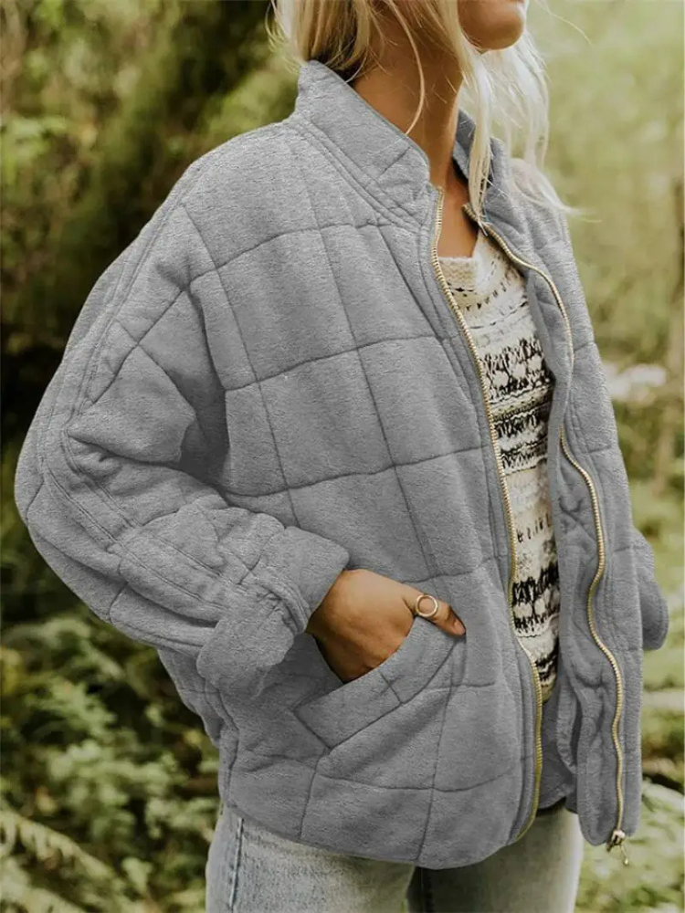 Frau in grauer Steppjacke, lässig über gemustertem Pullover, steht in natürlicher Umgebung. Modetrend Herbst, bequeme Outdoor-Bekleidung.