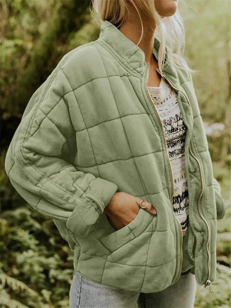 Blonde Frau in grüner Steppjacke mit Reißverschluss, lässig über gestreiftem Pullover und Jeans getragen, steht in natürlicher Waldumgebung.