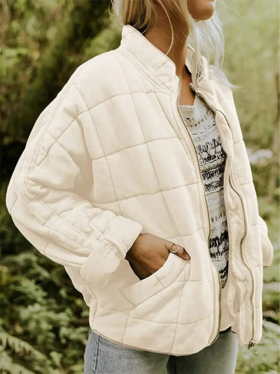 Frau in cremefarbener Steppjacke mit Reißverschluss, lässig über gestreiftem Pullover und Jeans, im Freien stehend. Modetrend Herbst 2023.