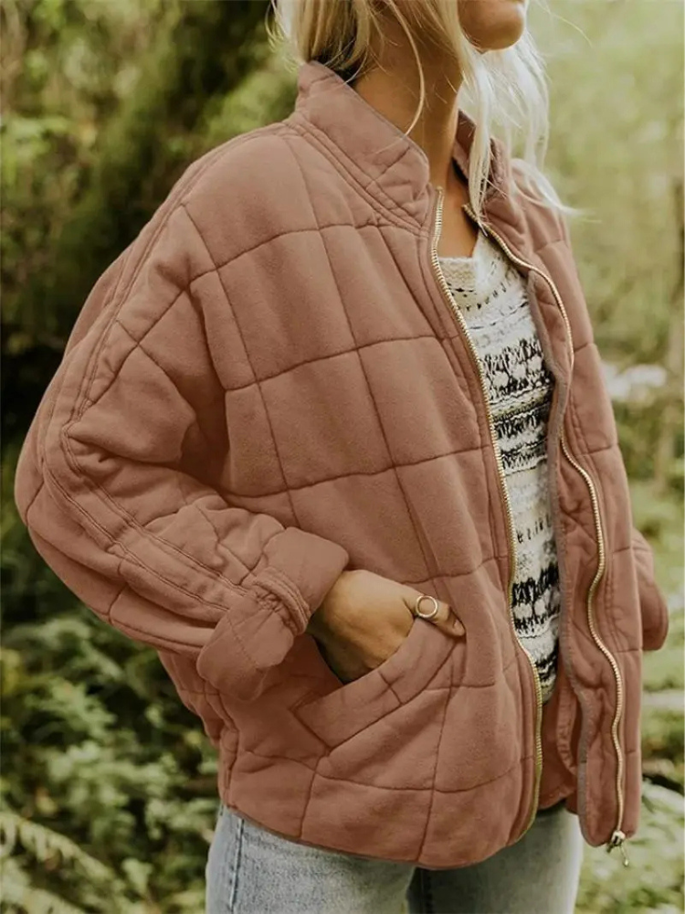 Frau in rosa Steppjacke mit Reißverschluss, lässig über gemustertem Pullover und Jeans getragen, steht in natürlicher Umgebung. Modetrend Herbst 2023.
