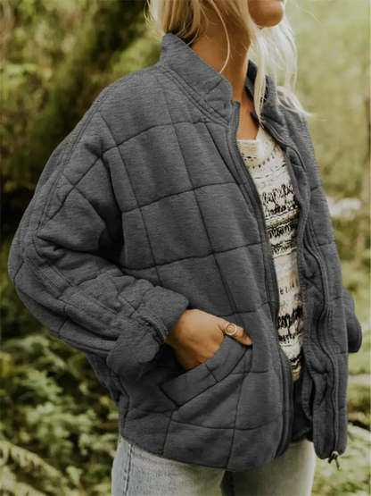 Frau in grauer Steppjacke, lässig über gemustertem Pullover, steht in natürlicher Umgebung. Modetrend Herbst, Outdoor-Bekleidung, Damenmode.