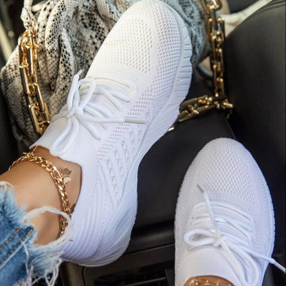 Weiße Sneaker mit Strickmuster, getragen von Person mit Jeans und goldenen Fußkettchen, neben einer Tasche mit Kettendetails im Auto.