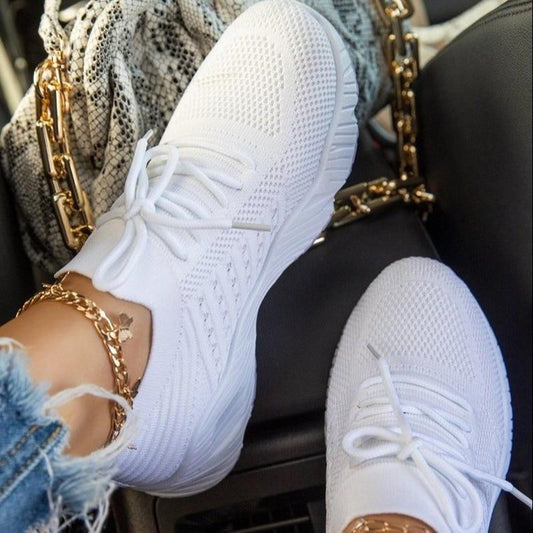 Weiße Sneaker mit Strickmuster, getragen von Person mit Jeans und goldenen Fußkettchen, neben einer Tasche mit Kettendetails im Auto.
