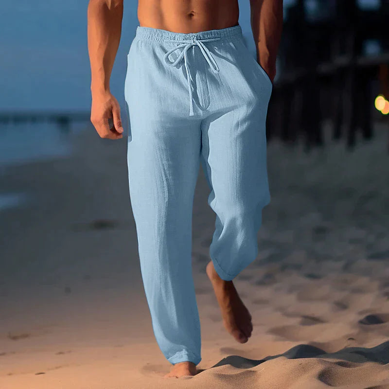 Mann in hellblauer Leinenhose spaziert barfuß am Strand entlang bei Sonnenuntergang. Sommermode, Strandkleidung, entspannter Stil.