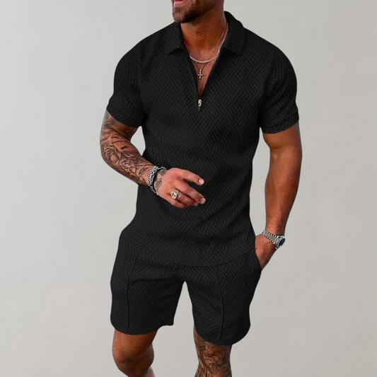 Mann in schwarzem, gemustertem Kurzarm-Overall, tätowierte Arme, trägt Schmuck, lässige Pose. Herrenmode, Sommeroutfit, trendiger Look.