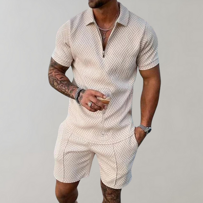 Mann in beigem, gemustertem Kurzarm-Poloshirt und Shorts, hält Getränk. Sommermode, Herrenbekleidung, lässiger Stil, tätowierter Arm.