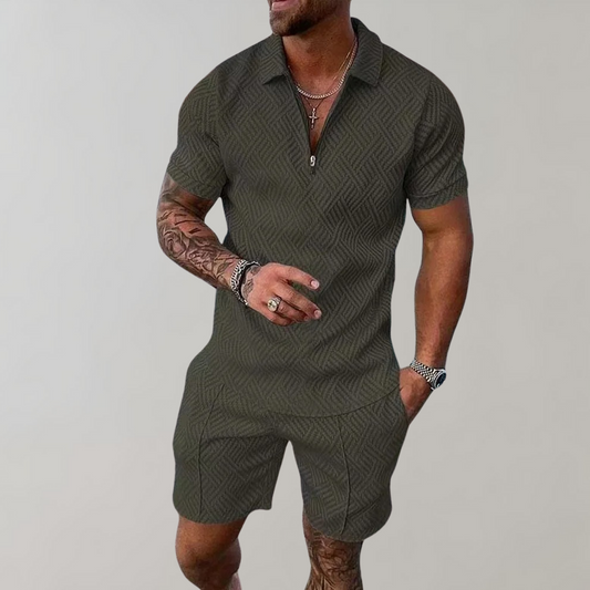 Mann in olivgrünem, gemustertem Kurzarm-Overall mit Reißverschluss, tätowierten Armen und Schmuck. Modischer Herren-Einteiler, Sommeroutfit.