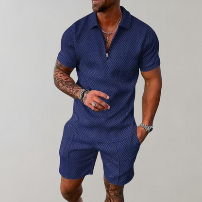 Mann in blauem, gemustertem Kurzarm-Overall, tätowierte Arme, trägt Schmuck, lässige Pose. Herrenmode, Sommeroutfit, trendiger Look.