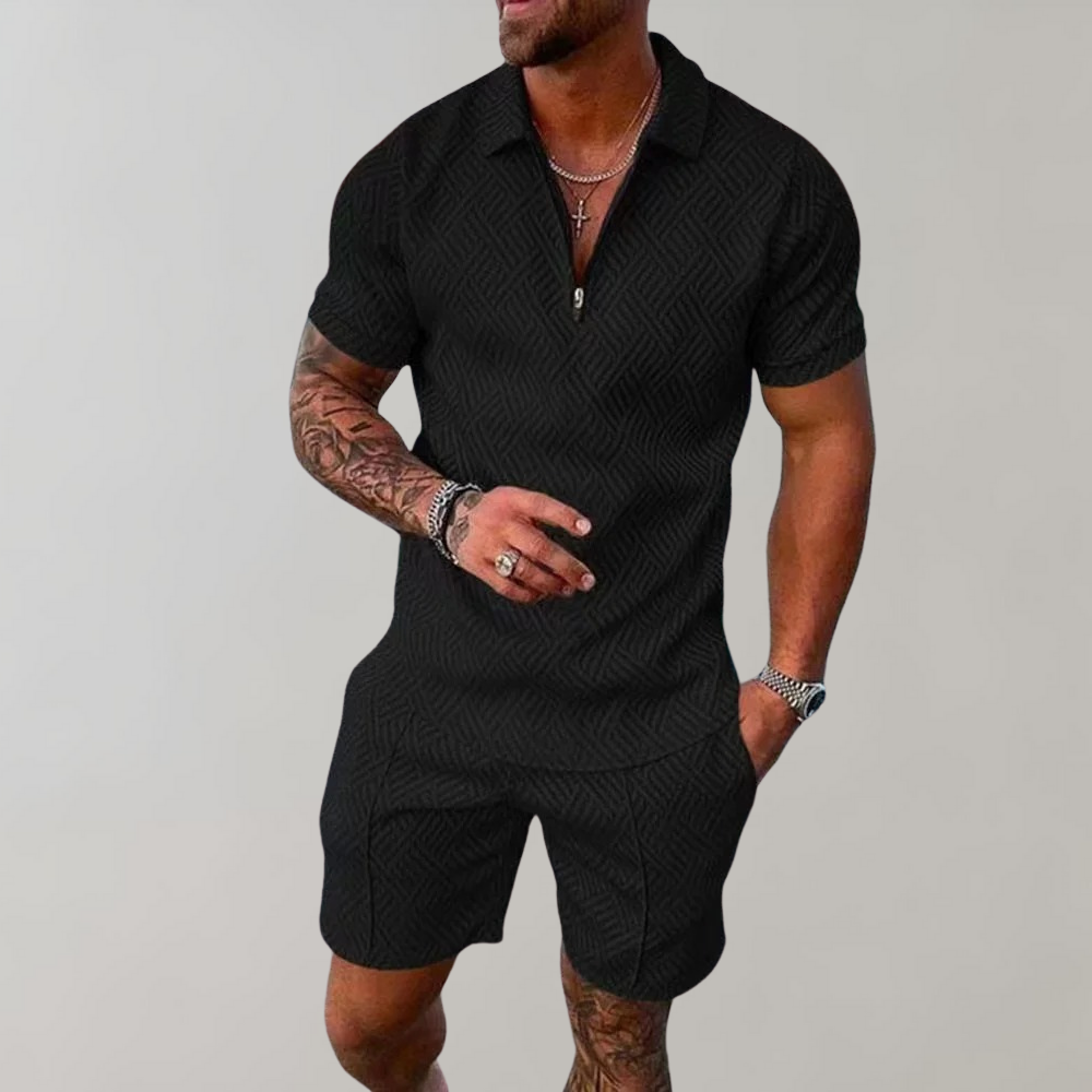 Schwarzer Herren-Shorts-Overall mit geometrischem Muster, getragen von muskulösem Mann mit Tattoos, silbernen Ketten und Armbändern vor grauem Hintergrund.