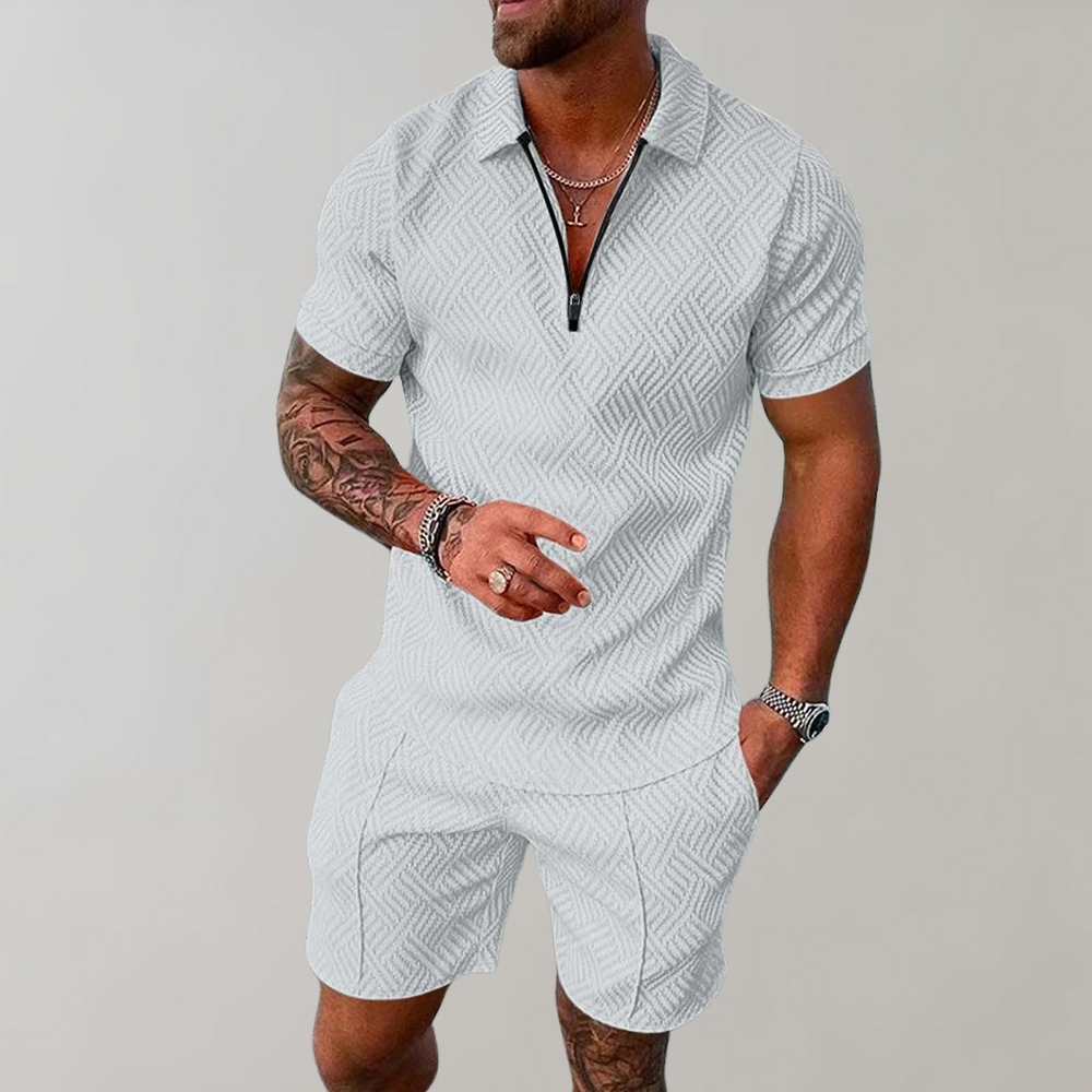 Mann in weißem, gemustertem Kurzarm-Poloshirt und Shorts, zeigt tätowierten Arm, trägt Armbanduhr und Kette. Sommermode, Herrenbekleidung.