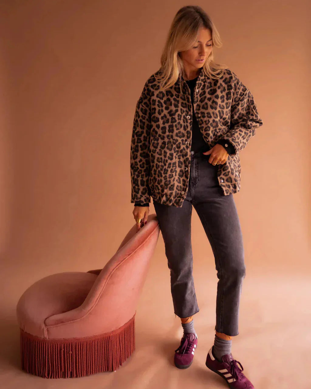 Frau in Leopardenjacke, grauer Jeans und lila Sneakers steht neben rosa Sessel. Modetrend, Herbstmode, stilvolle Damenbekleidung.