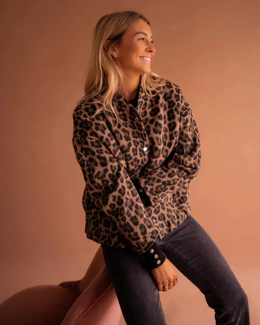 Blonde Frau in Leopardenmuster-Jacke, lächelnd, sitzt auf rosa Stuhl vor braunem Hintergrund. Modetrend, Herbstmode, Damenbekleidung.