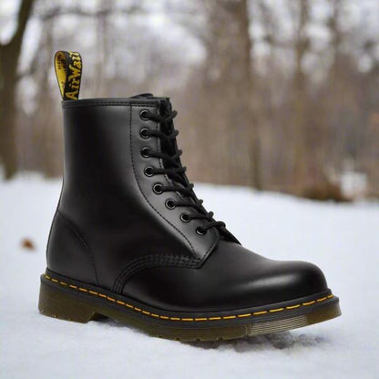 Schwarzer Lederstiefel mit gelber Naht und Schnürung auf schneebedecktem Boden im Wald. Robuster Winterstiefel, ideal für Outdoor-Aktivitäten.