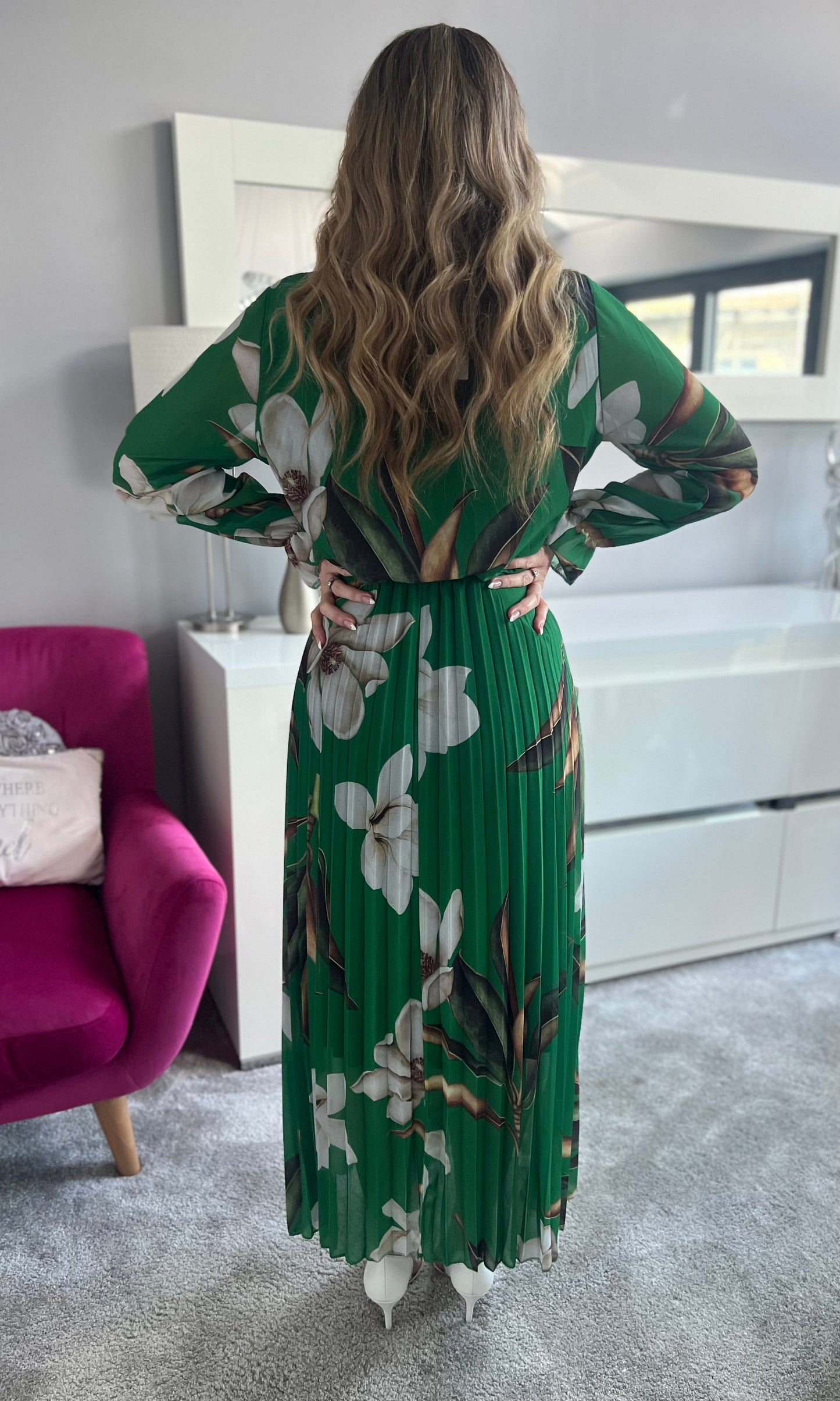 Frau in langem, grünem Kleid mit weißen Blumen, steht im modernen Wohnzimmer. Elegantes, florales Design, welliges Haar, stilvolle Einrichtung.